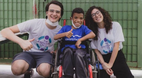 Imagem com dois voluntários do Friendship Circle e uma criança deficiente em cadeira de rodas.
