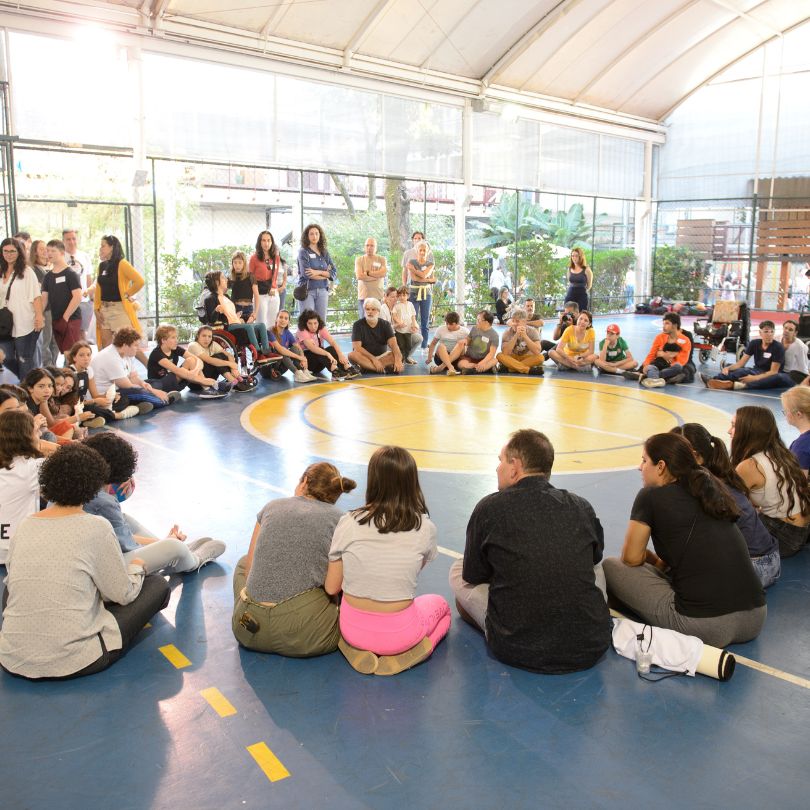 Grupo de pessoas sentadas em círculo em uma quadra, interagindo e compartilhando experiências em um ambiente colaborativo.