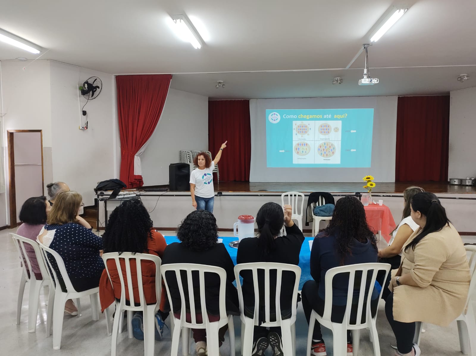 Uma mulher está de pé e aponta para um slide de apresentação diante de uma plateia sentada, em uma sala bem iluminada com paredes brancas e cortinas vermelhas.