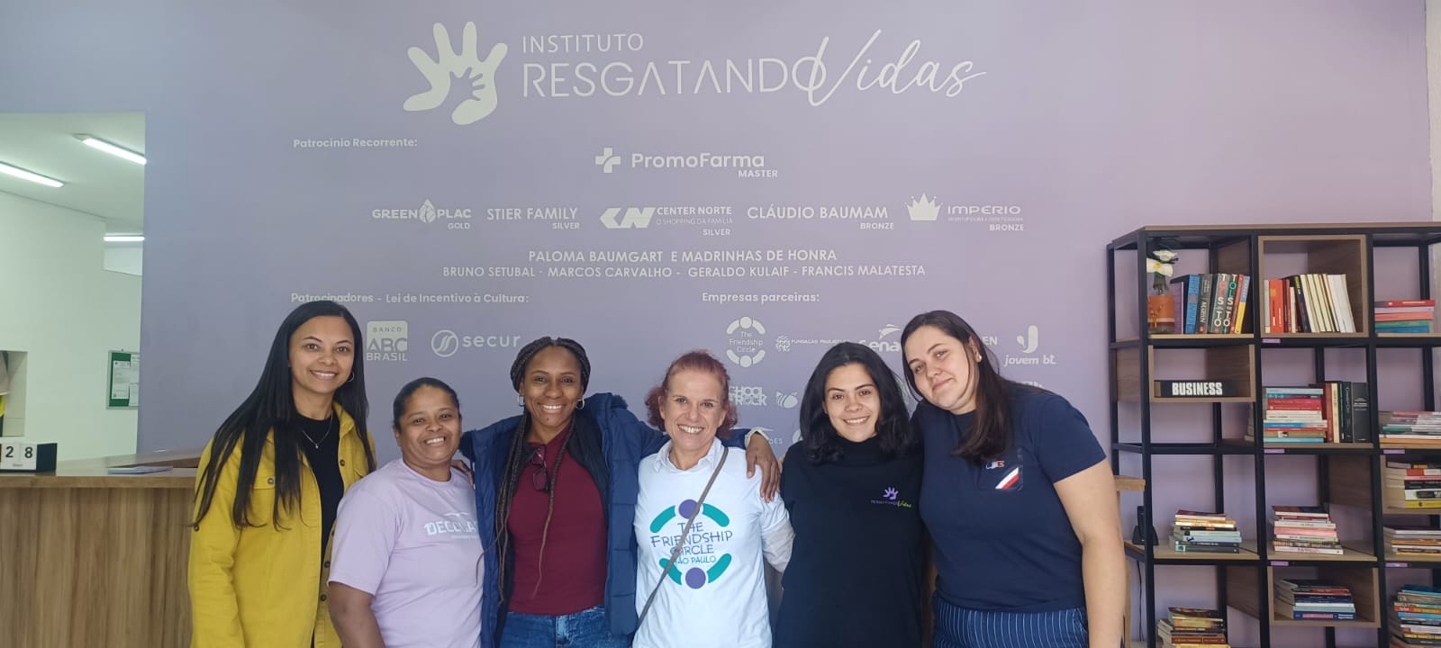 Seis mulheres estão de pé, sorrindo, diante de uma parede roxa com o logotipo do “Instituto Resgatando Vidas” e os nomes dos patrocinadores, ao lado de uma estante repleta de livros.
