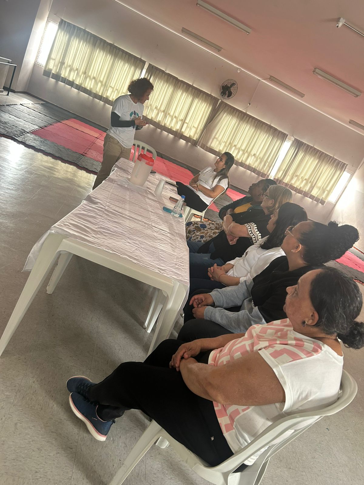 Uma pessoa está de pé e fala junto a uma mesa, diante de um grupo de pessoas sentadas, em uma sala com janelas grandes e tapetes vermelhos no chão.