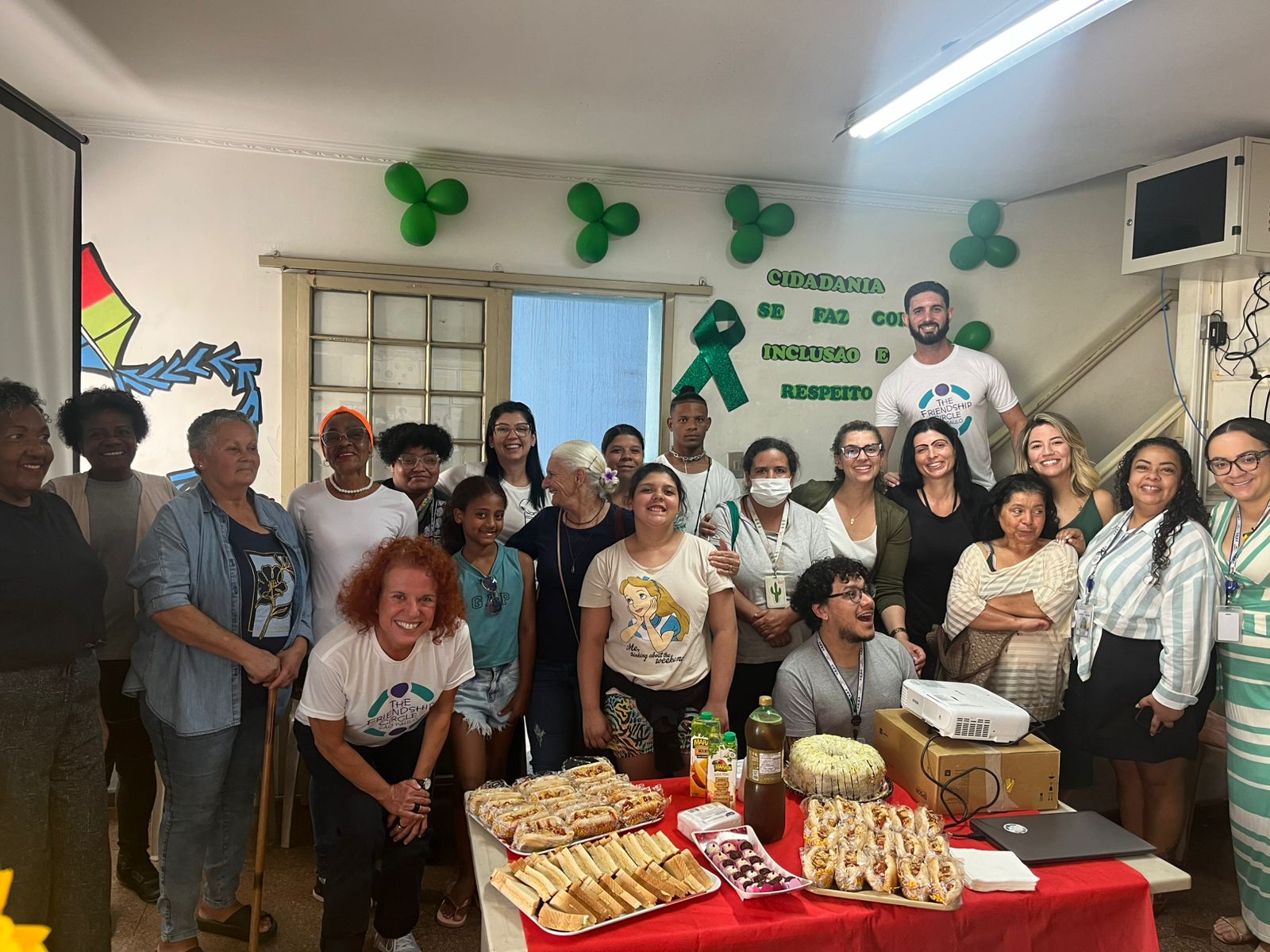 Um grupo diversificado de pessoas sorri para uma foto em grupo em uma sala decorada com fitas verdes na parede. Uma mesa à frente está repleta de comida e bebidas, o que sugere uma comemoração ou um evento comunitário.