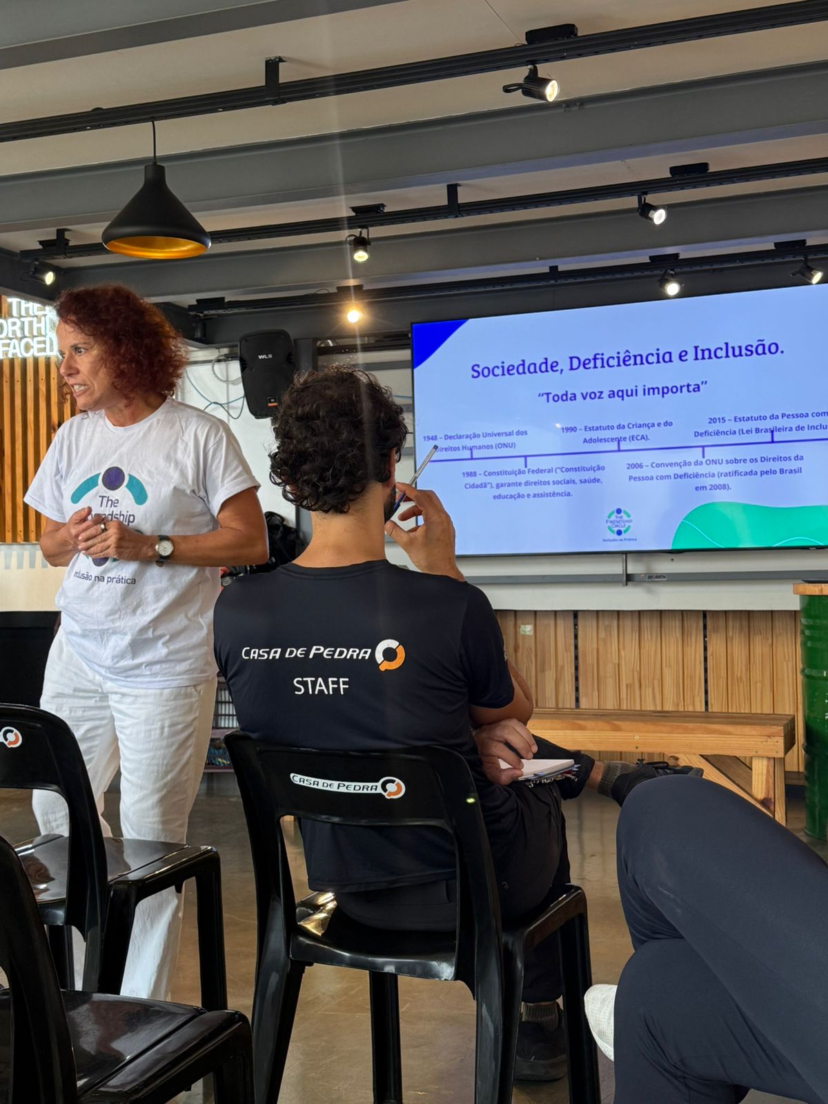 Uma mulher está de pé, falando diante de uma plateia sentada. Atrás dela, há um slide de apresentação com o título "Sociedade, Deficiência e Inclusão". O ambiente parece ser um espaço interno destinado a workshops ou seminários.