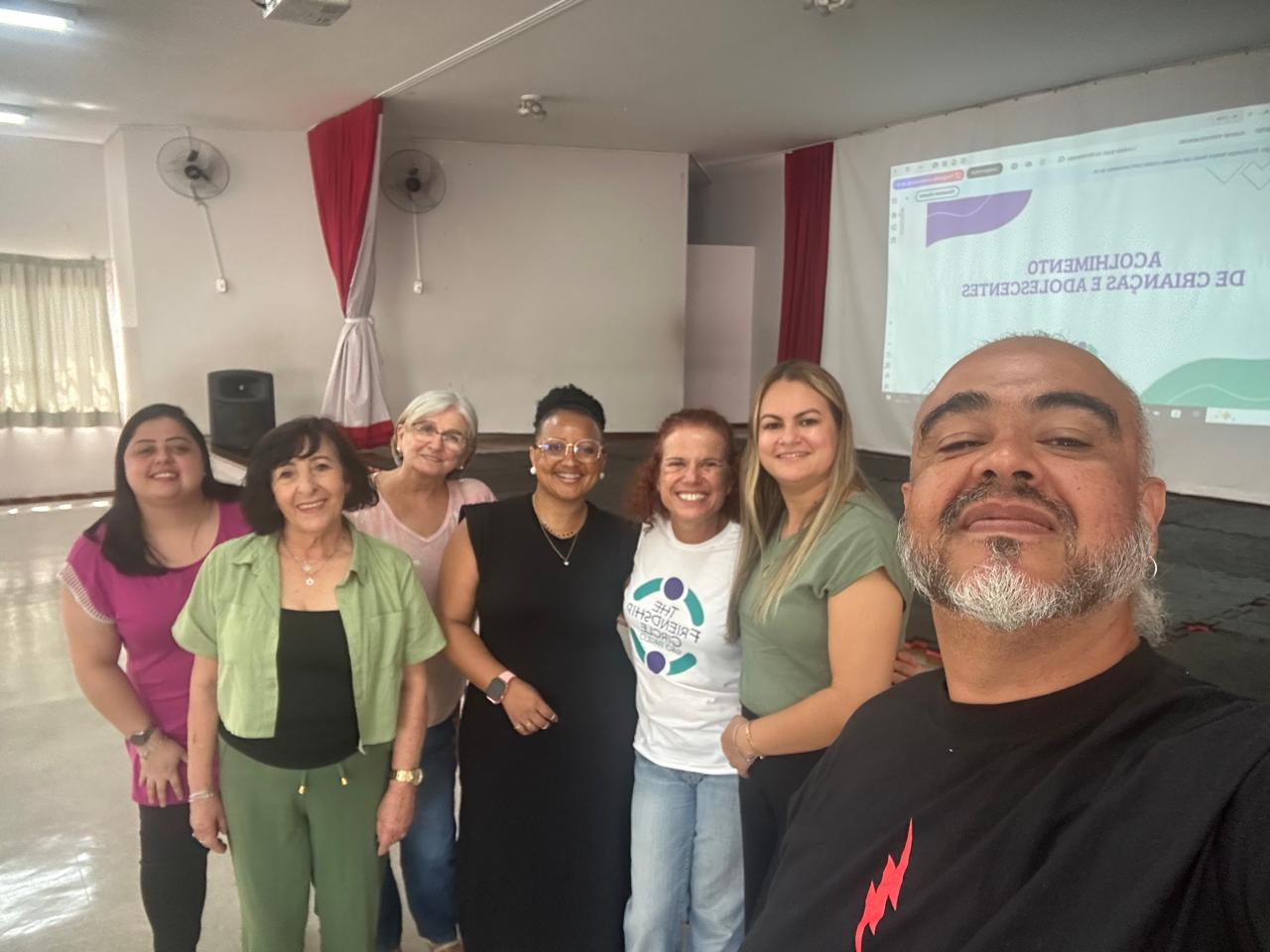 Sete pessoas estão juntas e sorridentes em um ambiente interno, com uma apresentação projetada em uma tela ao fundo. O grupo parece amigável e diverso, e a sala é bem iluminada, com paredes brancas e cortinas vermelhas.