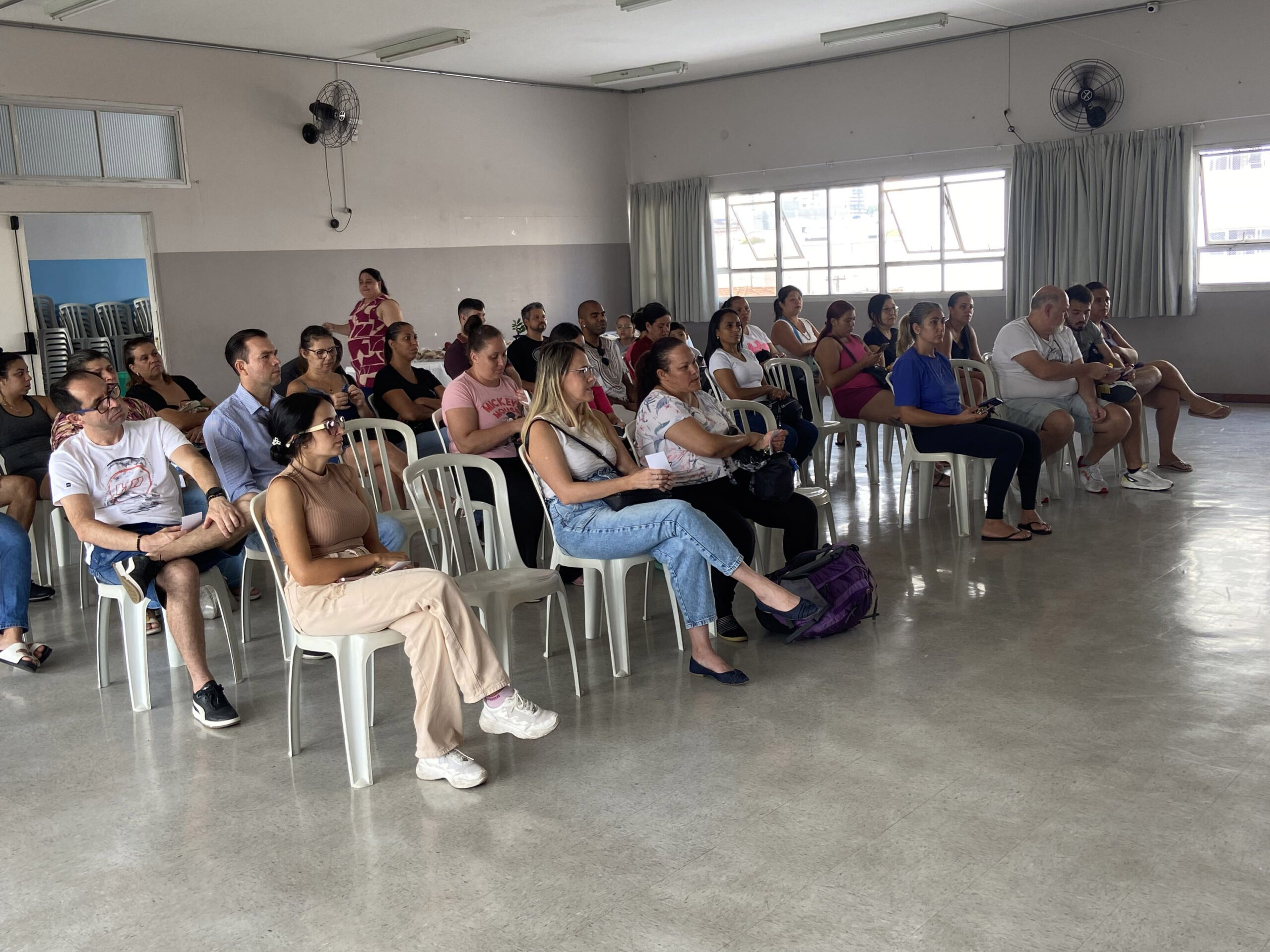 Um grupo de adultos está sentado em cadeiras de plástico brancas em uma sala bem iluminada, voltado atentamente para a frente, como se ouvisse uma apresentação ou participasse de uma reunião. A sala possui janelas grandes, paredes brancas e cortinas de cor clara.