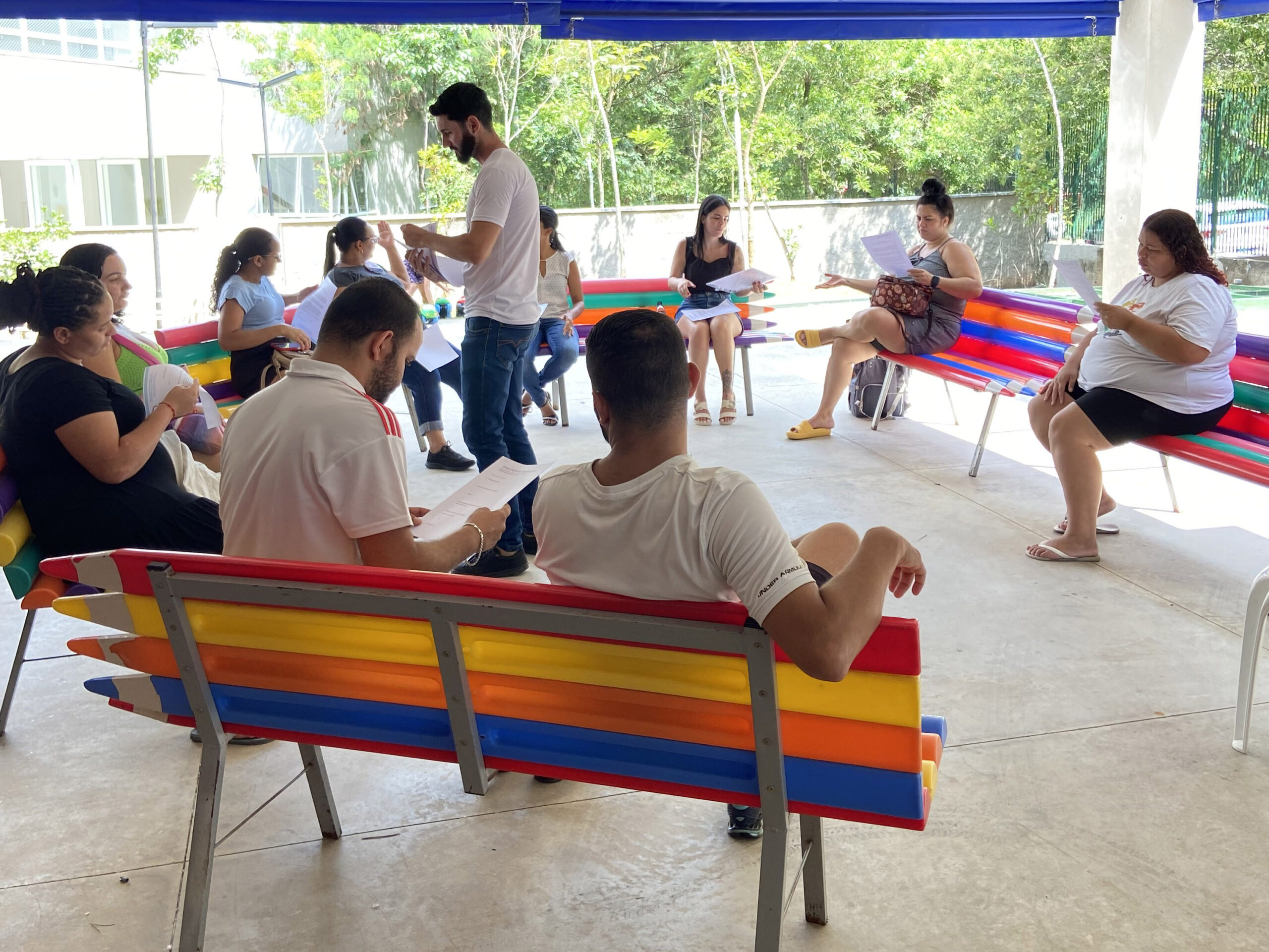 Um grupo de adultos está sentado em bancos coloridos em formato de lápis, lendo jornais e conversando, em uma área externa coberta, com árvores e prédios ao fundo.