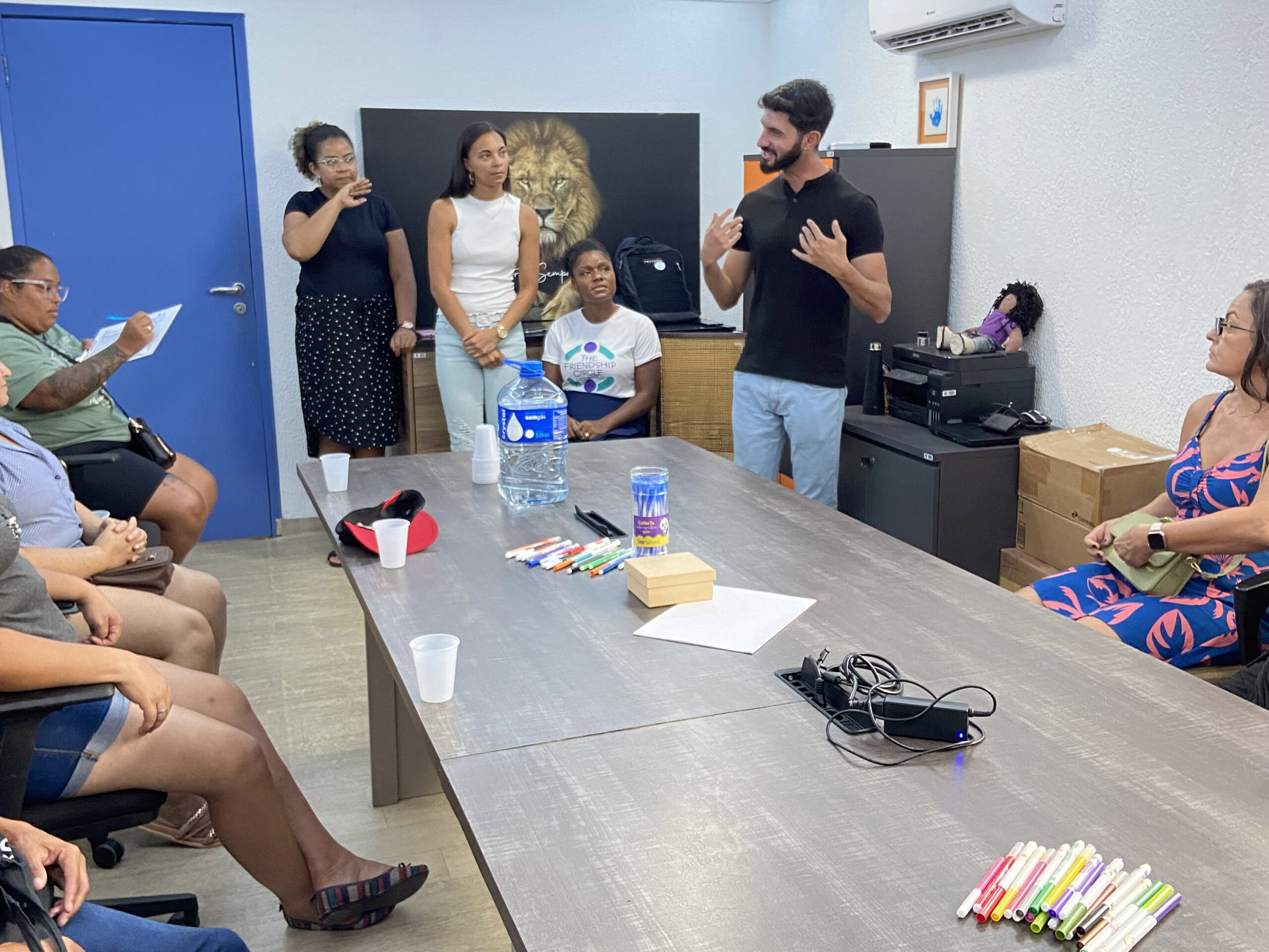 Um grupo de pessoas está reunido em um escritório, ao redor de uma mesa com marcadores e papéis. Um homem está falando, enquanto os outros ouvem e fazem anotações.