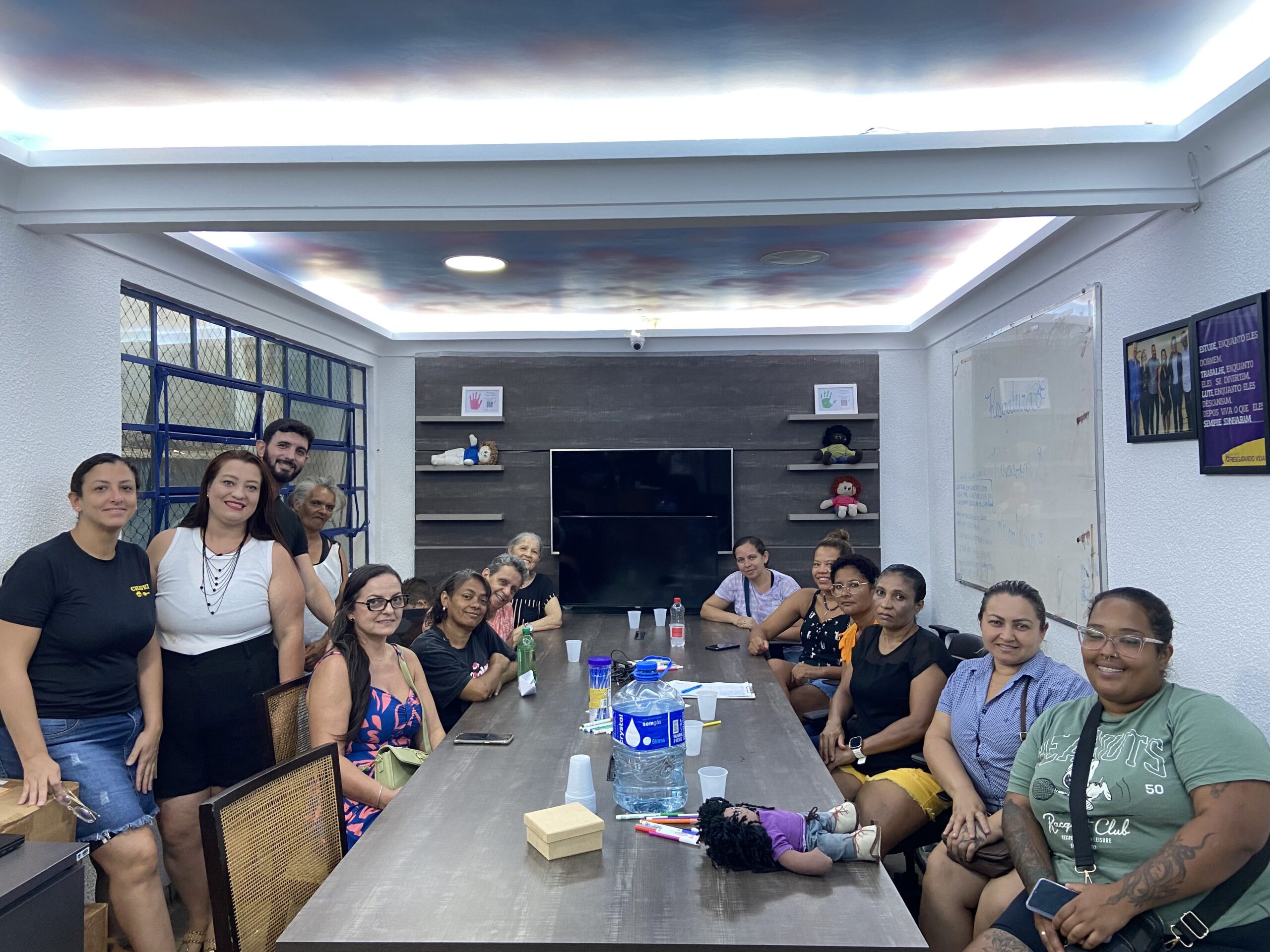 Um grupo de pessoas está sentado e em pé ao redor de uma mesa de conferência em uma sala de reuniões, sorrindo para a câmera. Há cadernos, bebidas e uma boneca sobre a mesa, com uma TV e um quadro branco ao fundo.